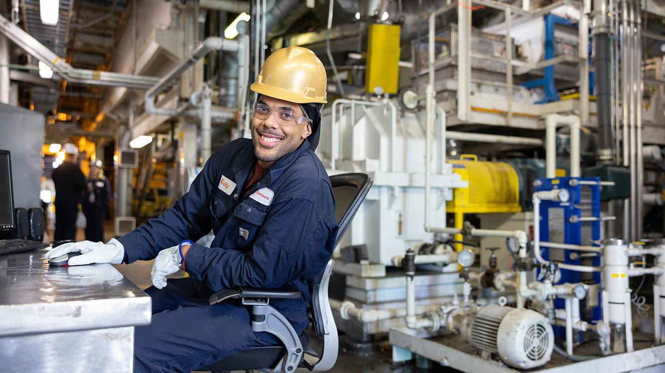 Man in hardhat and safety glasses sitting at a desk in a factory