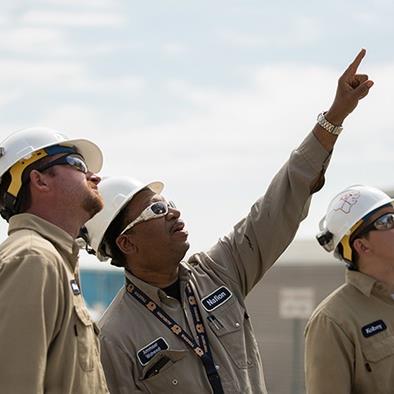 three men in white hardhats tan coveralls and sun glasses looking up at something the middle man is