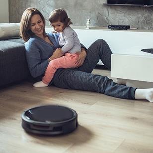 woman holding child on her lap both watching a robot vacuum