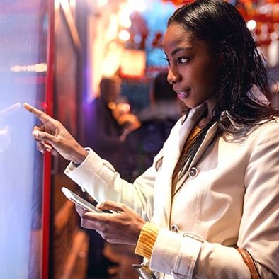 An image of a woman standing in front of a touchscreen holding a phone.