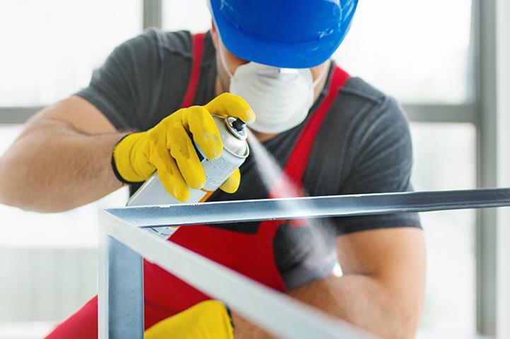 man in blue hardhat yellow gloves and mask spray painting a metal frame